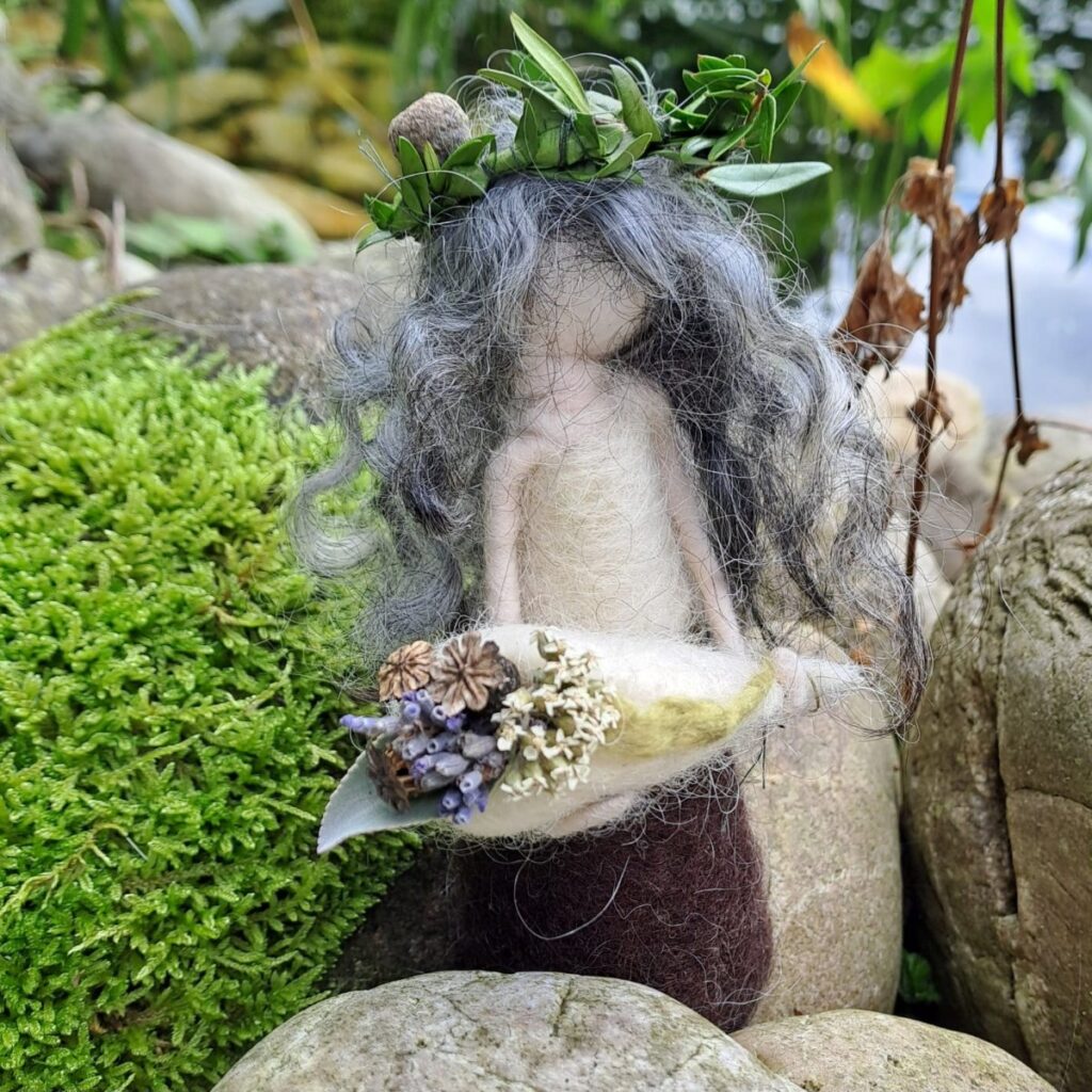 Gefilzte Figur mit Blätterkranz hält ein Blatt mit getrockneten Blüten, daneben leuchtend grünes Moos