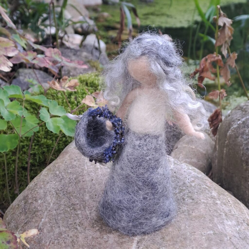 Gefilzte Figur in Grau mit hellem Haar, sie hält einen dunklen Kranz