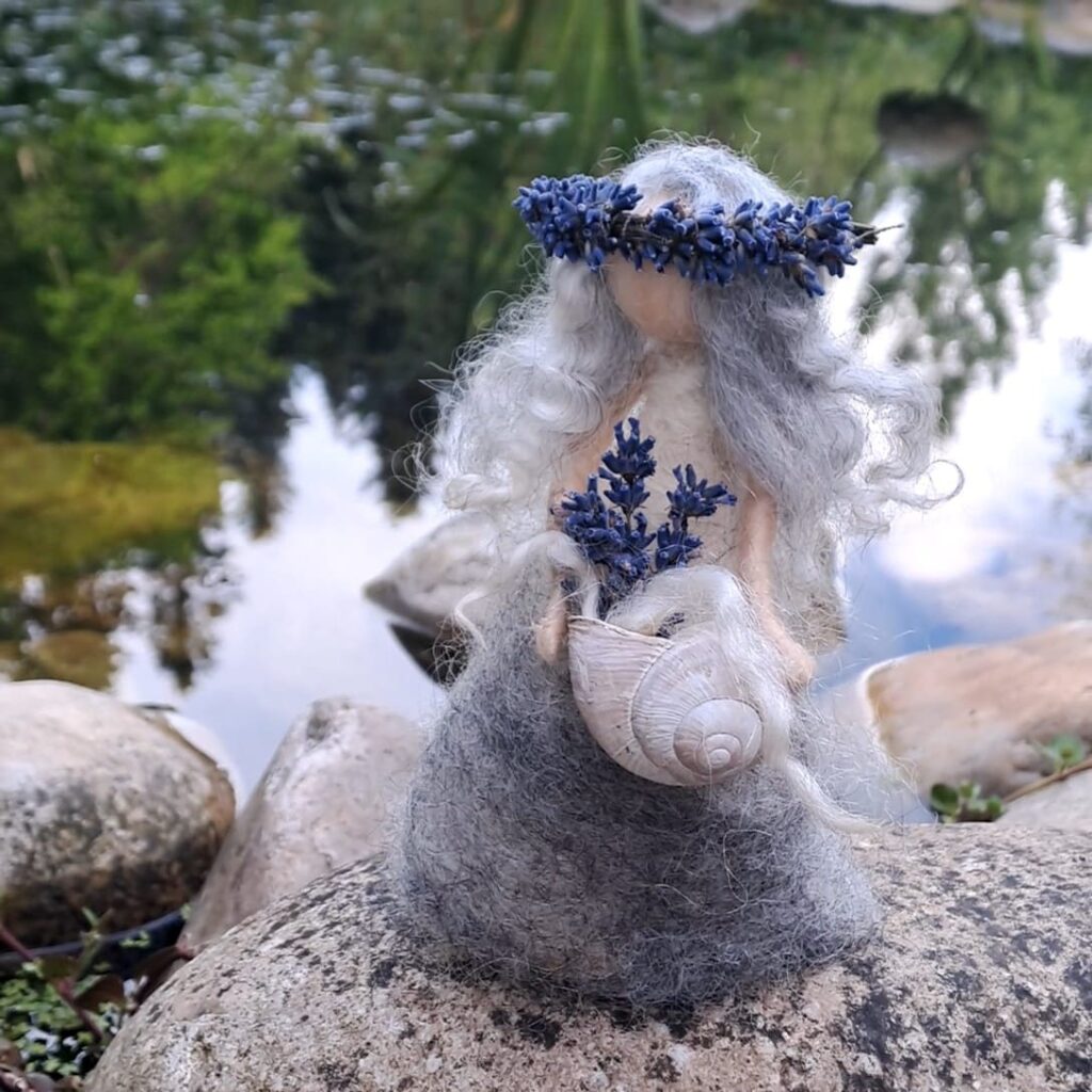 Gefilzte Figur mit Lavendelkranz und Schneckenhaus auf einem Stein am Wasser