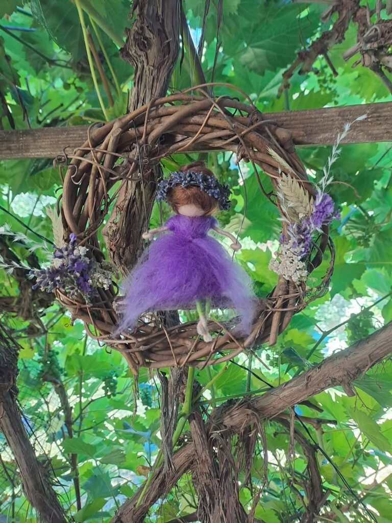 Gefilzte Figur in Lila mit Blumenkranz, hängend in einem Kranz aus Rebzweigen