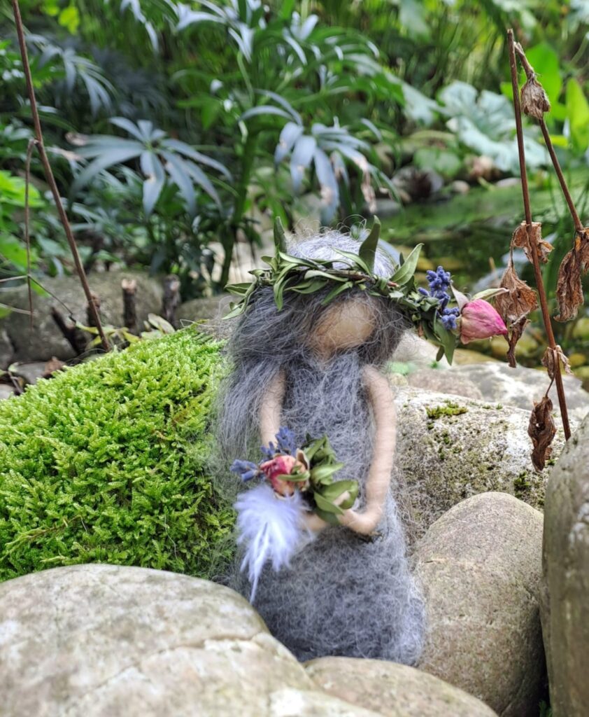 Gefilzte Figur mit Blätterkranz sitzt auf einem bemoosten Stein zwischen grünen Pflanzen