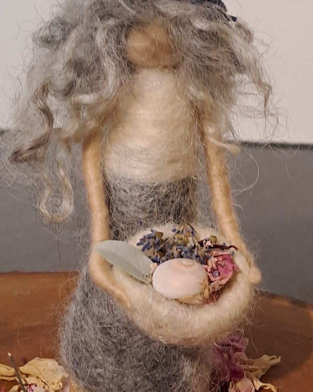 Gefilzte Figur mit Lavendelkranz hält eine Schale mit Schneckenhaus und getrockneten Blüten, auf einer Holzscheibe