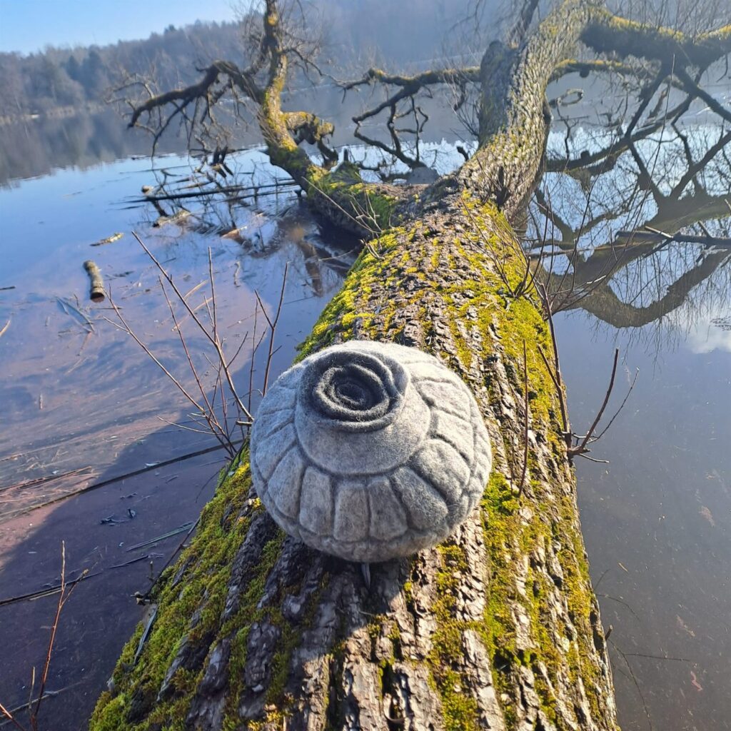 Gefilzte Urne mit Spiraldeckel auf einem bemoosten Baumstamm über dem Wasser