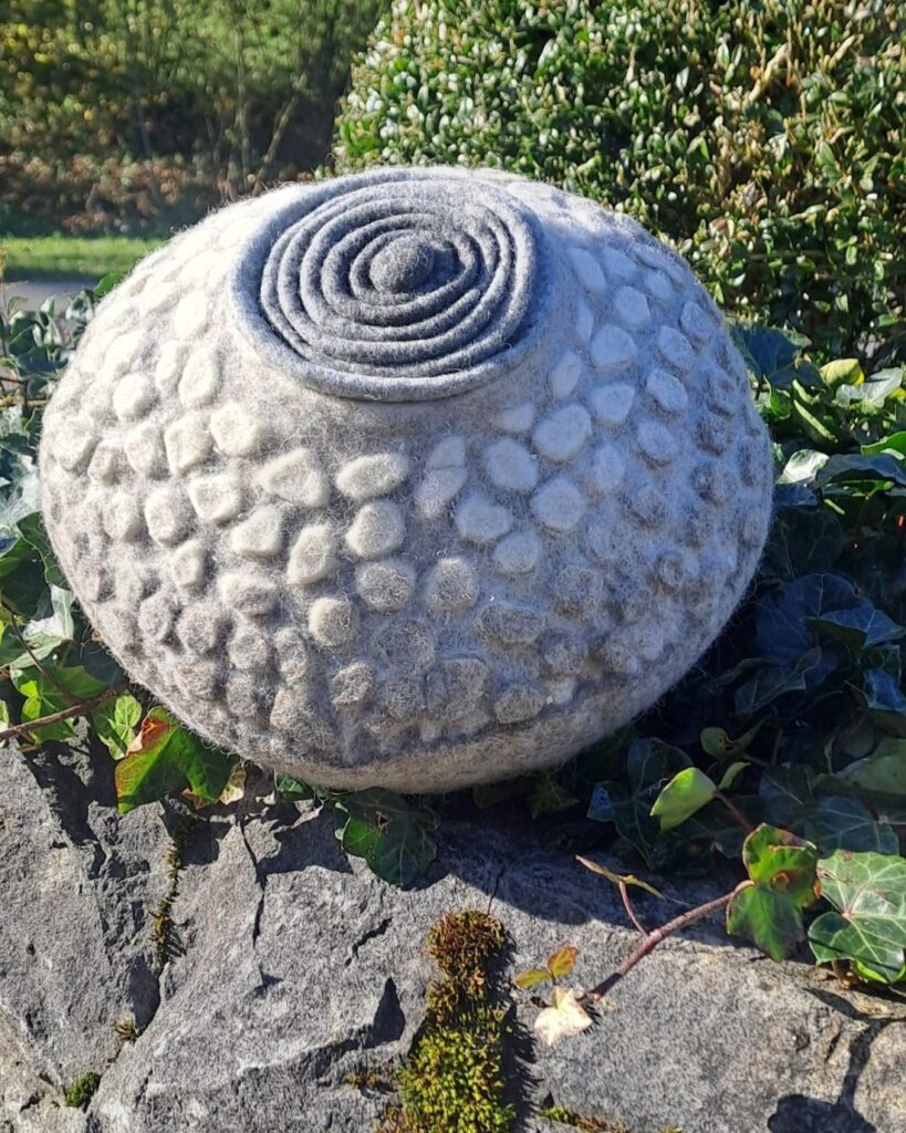 Graue gefilzte Urne mit genoppter Oberfläche und Spiraldeckel auf einem bemoosten Stein, umgeben von Efeu