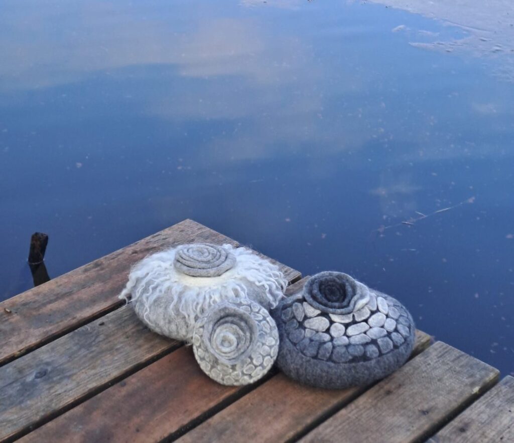 Gefilzte Urne mit gelockten Wollfransen und Spiraldeckel auf einem Holzsteg am See