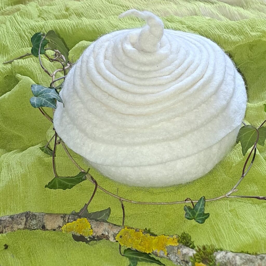 Weisse gefilzte Urne mit spiralförmigem Deckel auf grünem Filztuch, daneben Efeuranken und ein Ast mit gelber Flechte