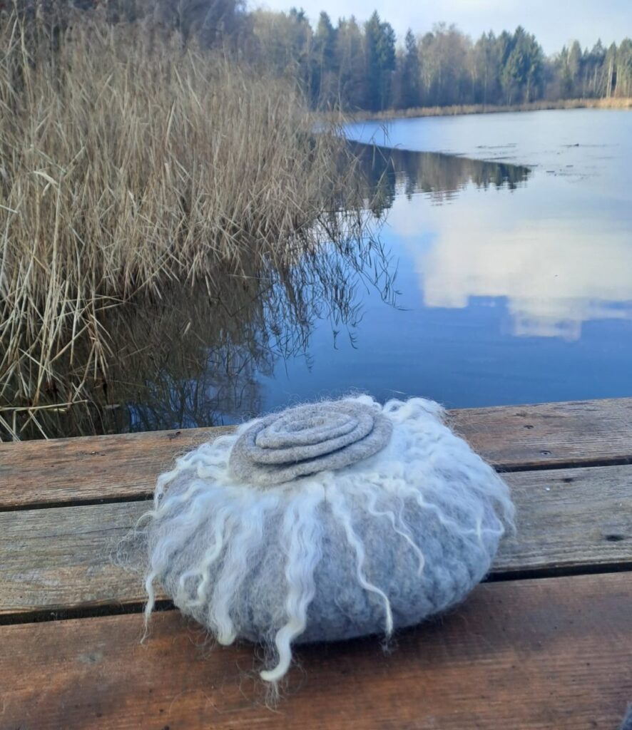 Weisse gefilzte Urne mit spitzem Spiraldeckel auf einem Holzsteg am See
