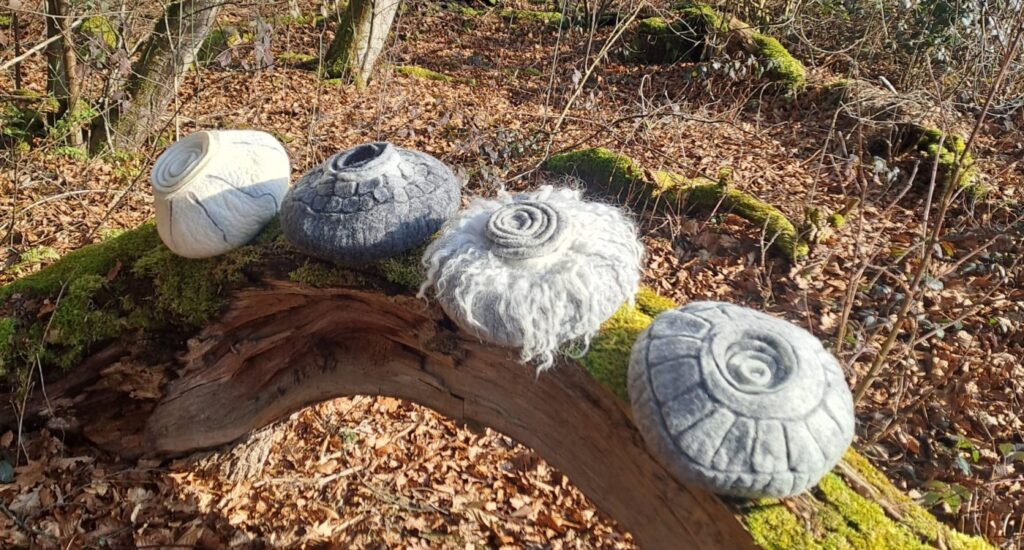 Vier verschiedene gefilzte Urnen aufgereiht auf einem bemoosten Ast im winterlichen Wald